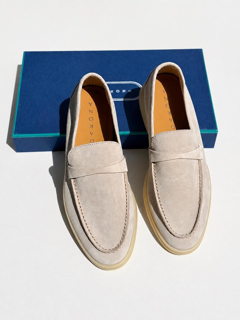 May include: A pair of beige suede loafers with a classic penny strap across the vamp, featuring light-colored rubber soles and tan leather insoles stamped with the brand name DARONA, positioned in front of a blue rectangular shoe box.