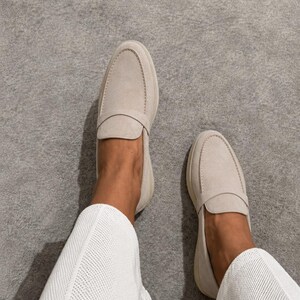 May include: A pair of light beige suede loafers featuring a rounded toe, a classic penny strap across the vamp, and a low-profile silhouette. The shoes are worn with white textured trousers, showcasing a clean and neutral aesthetic for everyday wear.