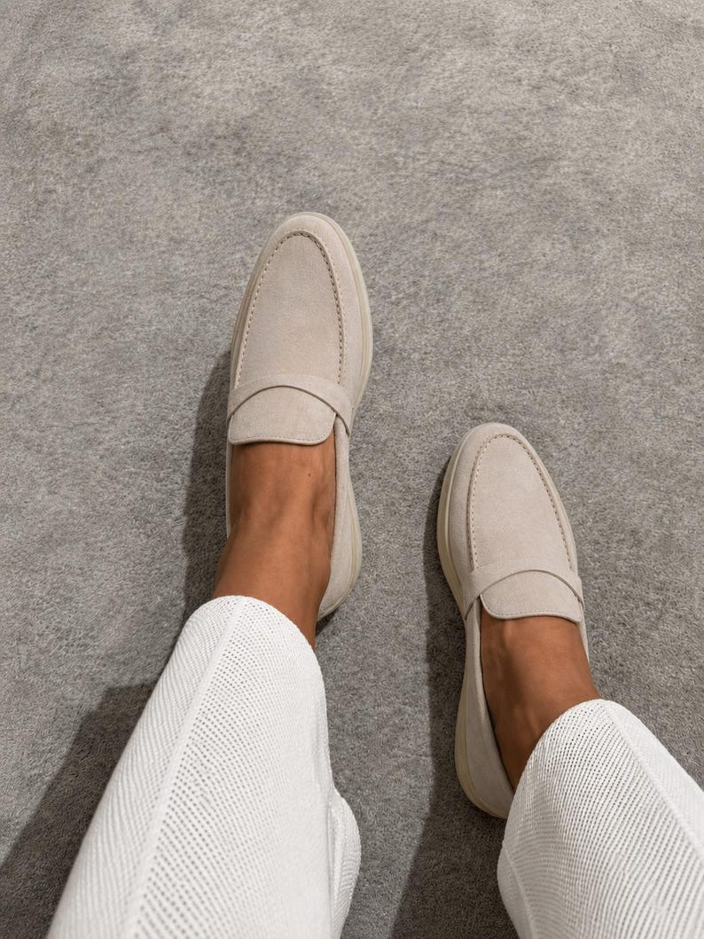 May include: A pair of light beige suede loafers featuring a rounded toe, a classic penny strap across the vamp, and a low-profile silhouette. The shoes are worn with white textured trousers, showcasing a clean and neutral aesthetic for everyday wear.