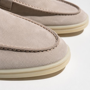 May include: A pair of light beige suede loafers featuring a moccasin-style stitched toe box, a decorative metallic buckle detail across the vamp, and a translucent rubber sole. The shoes have a soft matte texture and visible tonal stitching.