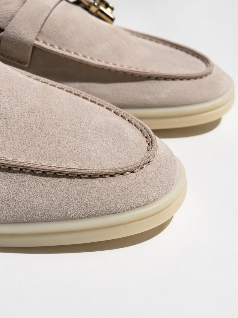 May include: A pair of light beige suede loafers featuring a moccasin-style stitched toe box, a decorative metallic buckle detail across the vamp, and a translucent rubber sole. The shoes have a soft matte texture and visible tonal stitching.