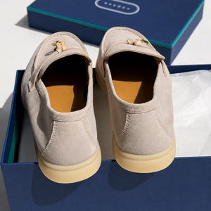 May include: A pair of beige suede loafers with a gold-toned metal tassel detail on the front. The shoes feature a tan interior lining and a thick, light-colored rubber sole. They are presented inside a dark blue rectangular cardboard box.