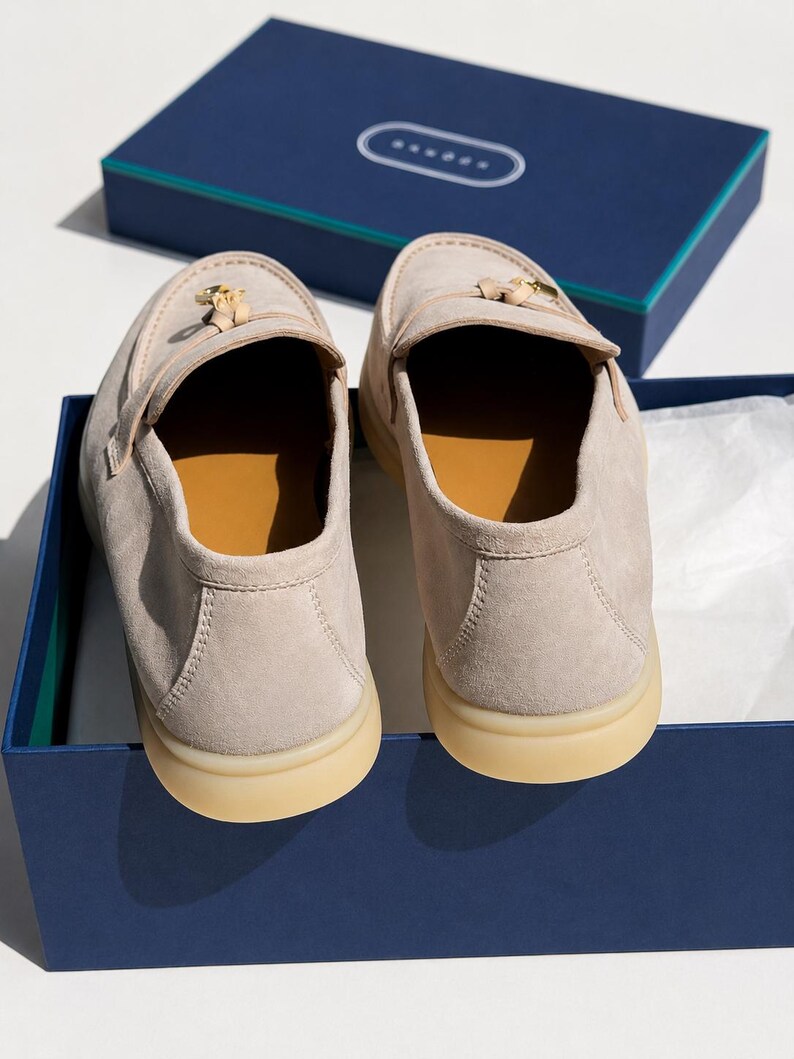 May include: A pair of beige suede loafers with a gold-toned metal tassel detail on the front. The shoes feature a tan interior lining and a thick, light-colored rubber sole. They are presented inside a dark blue rectangular cardboard box.