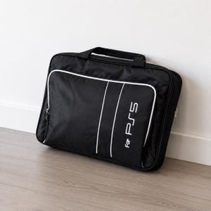 Op de afbeelding: Zwarte draagtas voor een PS5-console. Witte rand en verticale witte strepen. De tekst "For PS5" is wit gedrukt. Met handvat en ritssluiting. Ontworpen om een PS5-console te beschermen en te vervoeren.