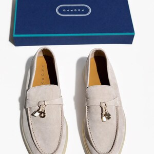 May include: A pair of light beige suede loafers featuring a decorative strap with a small gold-toned padlock and ring charm. The interior soles are tan and labeled with the brand name ARONA. A matching dark blue rectangular box is included.