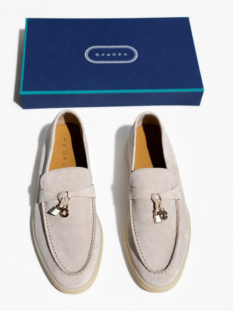 May include: A pair of light beige suede loafers featuring a decorative strap with a small gold-toned padlock and ring charm. The interior soles are tan and labeled with the brand name ARONA. A matching dark blue rectangular box is included.