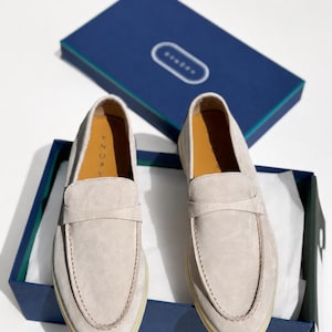 May include: A pair of light beige suede loafers with a classic penny strap design, featuring a rounded toe and a flat rubber sole. The shoes are presented inside a dark blue rectangular box with a matching lid, displaying the brand name AZORA.