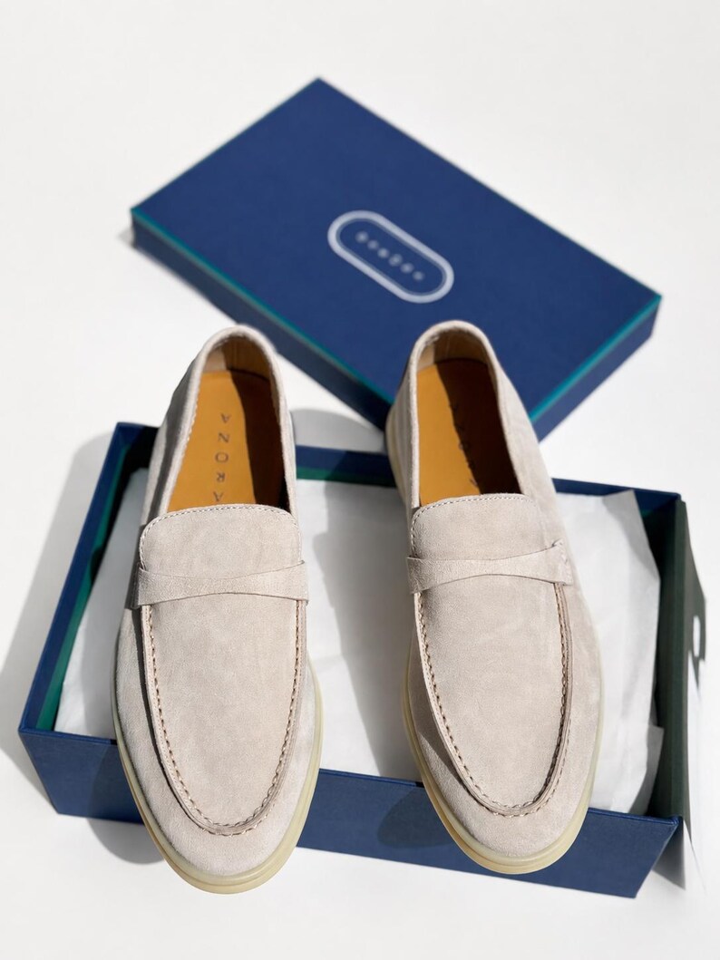 May include: A pair of light beige suede loafers with a classic penny strap design, featuring a rounded toe and a flat rubber sole. The shoes are presented inside a dark blue rectangular box with a matching lid, displaying the brand name AZORA.