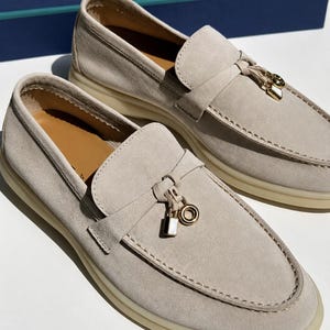 May include: A pair of beige suede loafers featuring a gold-toned metal charm on the vamp, consisting of a small padlock and a circular ring. The shoes have a thick, light-colored rubber sole and a tan interior lining, showcasing visible stitching.