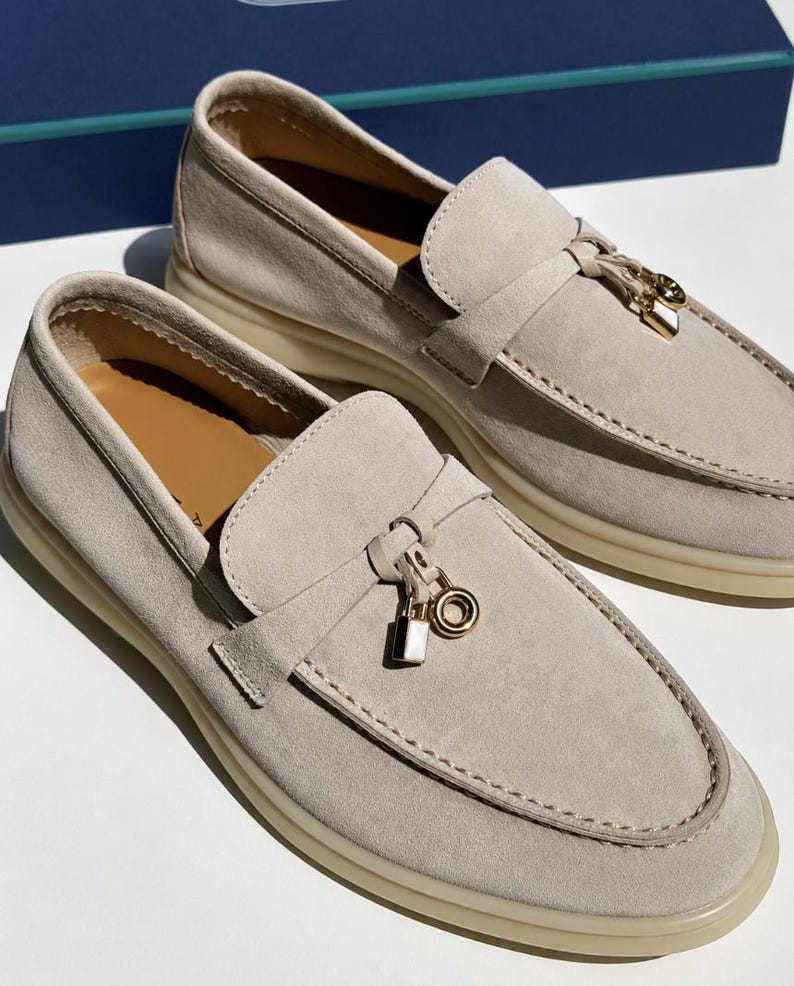 May include: A pair of beige suede loafers featuring a gold-toned metal charm on the vamp, consisting of a small padlock and a circular ring. The shoes have a thick, light-colored rubber sole and a tan interior lining, showcasing visible stitching.