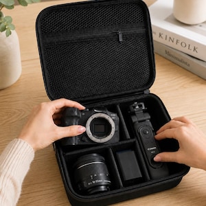 K&ouml;nnte beinhalten: Schwarze Kameratasche mit einer Canon EOS R7 Kamera, Objektiv, Akku und Fernbedienung. Die Tasche hat eine Netztasche auf der Innenseite des Deckels. Die Tasche befindet sich auf einer Holzoberfl&auml;che.