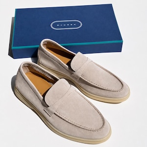 May include: A pair of light beige suede loafers with a moccasin-style stitched toe, tan leather insoles, and thick cream-colored rubber soles. The shoes are positioned in front of a rectangular navy blue box featuring the white text DAKONA.