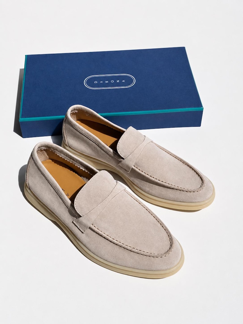 May include: A pair of light beige suede loafers with a moccasin-style stitched toe, tan leather insoles, and thick cream-colored rubber soles. The shoes are positioned in front of a rectangular navy blue box featuring the white text DAKONA.
