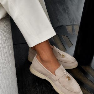 May include: A pair of beige suede loafers featuring a moccasin-style stitched toe, a flat rubber sole, and a decorative gold-toned metal charm detail attached to the front strap. The shoes are worn with white trousers.