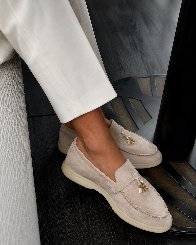 May include: A pair of beige suede loafers featuring a moccasin-style stitched toe, a flat rubber sole, and a decorative gold-toned metal charm detail attached to the front strap. The shoes are worn with white trousers.