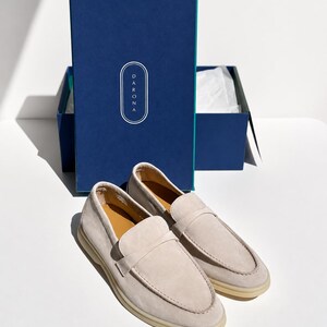 May include: A pair of light beige suede loafers with a rounded toe and a strap across the vamp, positioned in front of a dark blue rectangular box featuring the white text DARONA inside an oval outline and a teal interior edge.