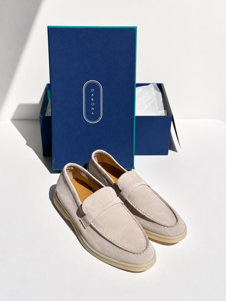 May include: A pair of light beige suede loafers with a rounded toe and a strap across the vamp, positioned in front of a dark blue rectangular box featuring the white text DARONA inside an oval outline and a teal interior edge.