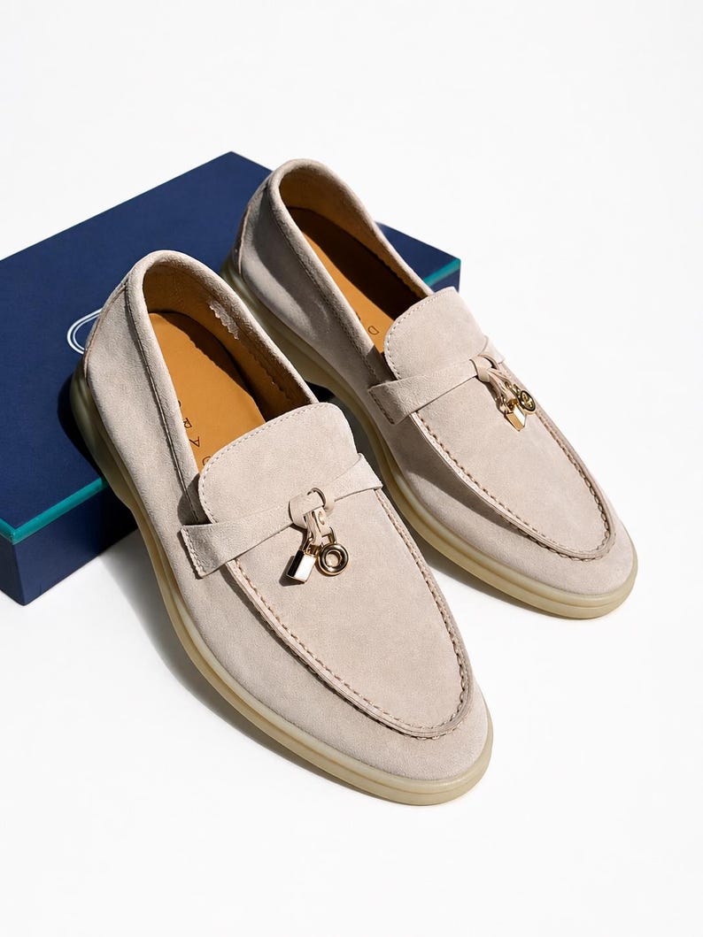 May include: A pair of beige suede loafers featuring a moccasin-style toe, a horizontal strap across the vamp, and a gold-toned metal charm consisting of a small padlock and a circular ring. The shoes have a light-colored rubber sole and tan interior.