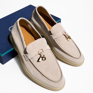 May include: A pair of beige suede loafers featuring a moccasin-style toe, a horizontal strap across the vamp, and a gold-toned metal charm consisting of a small padlock and a circular ring. The shoes have a light-colored rubber sole and tan interior.