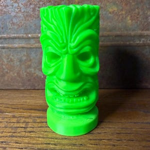 May include: A bright green tiki statue with a carved face, featuring large eyes, a wide nose, and a grinning mouth. The tiki is cylindrical with a flat base and is set against a rustic, weathered background.
