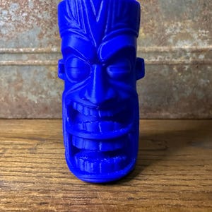 May include: A vibrant blue Tiki-style face sculpture with a wide, toothy grin, prominent eyes, and ear-like protrusions. The sculpture is made of a solid material and stands upright on a wooden surface, showcasing a detailed, carved design.