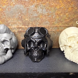 May include: Three decorative skull figurines. One is silver with a pompadour and beard, the second is black with goggles, and the third is off-white with a beard. They are displayed on a dark surface against a rusty metal background.