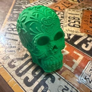 May include: A vibrant green decorative skull with intricate floral patterns. The skull has hollow eye sockets and a detailed nose and mouth. This unique piece is a Day of the Dead-themed item.