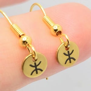 Puede incluir: Un par de pendientes dorados. Cada pendiente presenta un pequeño dije redondo con un símbolo negro. Los pendientes tienen un diseño de gancho para orejas perforadas. Los pendientes están sostenidos por un dedo.