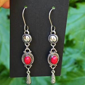 Puede incluir: Un par de pendientes colgantes plateados con piedras rojas de forma ovalada. Los pendientes tienen un diseño detallado con un dije en forma de lágrima en la parte inferior. Se muestran sobre un fondo negro con un telón de fondo verde y frondoso.