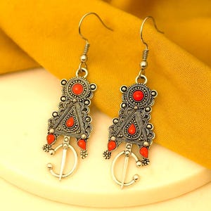 Puede incluir: Un par de pendientes plateados con un diseño geométrico y detalles rojos. Los pendientes tienen forma triangular con una parte superior circular y cuentas rojas colgantes. El fondo es una tela amarilla.