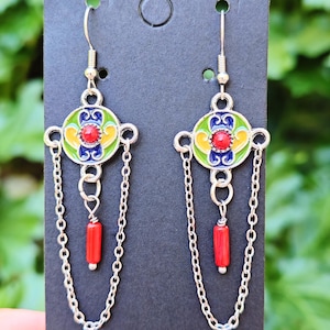 Puede incluir: Un par de pendientes plateados con un diseño floral colorido. Cada pendiente presenta una piedra central roja, rodeada de esmalte amarillo, verde y azul, con cuentas rojas colgantes y detalles de cadena.
