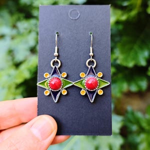 Puede incluir: Un par de pendientes colgantes plateados. Cada pendiente presenta una piedra central roja circular, rodeada por un diseño de estrella verde, azul y amarilla. Los pendientes se muestran en una tarjeta negra sobre un fondo verde borroso.