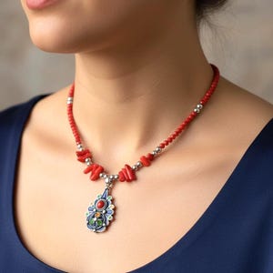 Puede incluir: Un collar de coral rojo con un colgante de plata. El colgante tiene un diseño de esmalte colorido con detalles en azul, verde, rojo y amarillo. El collar lo lleva una persona con una blusa azul oscuro.