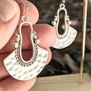 Puede incluir: Pendientes colgantes plateados con textura martillada y diseño en forma de abanico. Los pendientes presentan una forma de lágrima abierta en la parte superior, adornada con pequeños detalles de cuentas. Los pendientes son sostenidos por una mano.