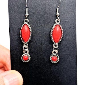 Puede incluir: Un par de pendientes plateados con detalles de piedras rojas ovaladas y redondas. Los pendientes tienen cierre de gancho y un borde decorativo con cuentas alrededor de las piedras rojas. Se muestran sobre fondo negro.