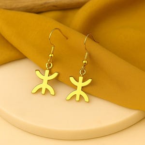 Puede incluir: Un par de pendientes dorados. Cada pendiente presenta un símbolo estilizado con una línea vertical central y brazos horizontales. Los pendientes están suspendidos de un gancho simple. El fondo es una tela de color amarillo mostaza.