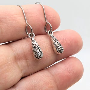 Puede incluir: Un par de pendientes colgantes plateados. Cada pendiente presenta un colgante en forma de lágrima con un diseño ornamental y arremolinado. Los pendientes tienen un cierre de gancho sencillo.