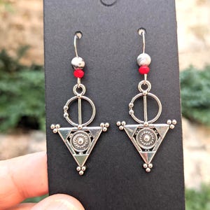 Puede incluir: Pendientes geométricos plateados con base triangular y detalles circulares. Los pendientes tienen cuentas rojas y blancas y se muestran en una tarjeta negra. El diseño es intrincado y detallado, con una estética bohemia.