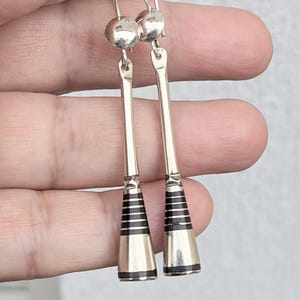 May include: Silver-toned dangle earrings. Each earring features a small ball at the top, a long, slender bar, and a cone-shaped bottom with black horizontal stripes. The earrings are held up by a silver hook.