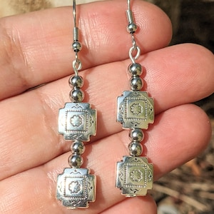 Puede incluir: Un par de pendientes de plata con un diseño del suroeste. Cada pendiente presenta un colgante cuadrado decorativo con un centro floral y detalles de cuentas. Los pendientes están suspendidos de un gancho plateado.
