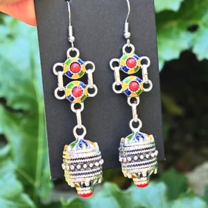 Puede incluir: Un par de pendientes colgantes plateados. Cada pendiente presenta un diseño geométrico colorido con detalles en rojo, amarillo, azul y verde. Los pendientes tienen una parte inferior texturizada detallada con una cuenta roja.