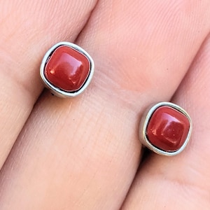 Puede incluir: Un par de pendientes de botón cuadrados. Cada pendiente presenta una piedra pulida de color rojo anaranjado engastada en un bisel de metal plateado. Los pendientes son pequeños y ligeros, adecuados para el uso diario.
