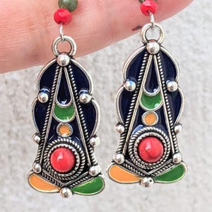 Puede incluir: Un par de pendientes colgantes ornamentados. Cada pendiente presenta un diseño esmaltado azul marino con detalles verdes, naranjas y rojos. Los pendientes tienen un acabado metálico plateado y están adornados con pequeñas cuentas.