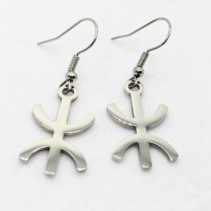 Puede incluir: Un par de pendientes de ancla de color plateado. Cada pendiente presenta un gancho, un resorte en espiral y un colgante de ancla estilizado. El diseño del ancla tiene una barra vertical con una parte superior curva y tres puntas curvas en la parte inferior.