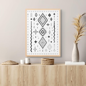 May include: A framed monochrome geometric print in a light wood frame. The artwork showcases diamond, cross, and zigzag patterns. The print is displayed on a wooden cabinet, alongside a vase and other decorative items.