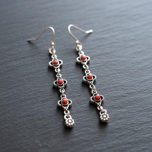 Puede incluir: Pendientes colgantes plateados con diseño floral y pequeñas cuentas rojas. Los pendientes tienen cierre de gancho y un elemento floral decorativo en la parte inferior. Se muestran sobre una superficie gris oscuro.