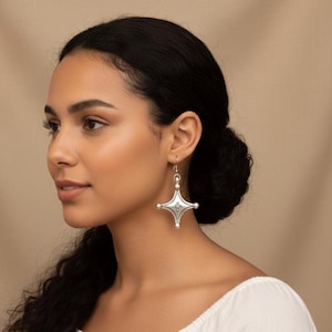 Puede incluir: Pendientes plateados en forma de cruz con detalles intrincados. Los pendientes presentan un diseño central con pequeños puntos en relieve y están suspendidos de un gancho simple. Una pieza llamativa.