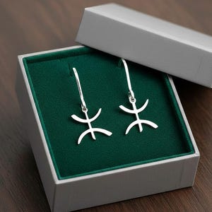 Puede incluir: Un par de pendientes plateados con un diseño estilizado, presentados en una caja gris con interior de terciopelo verde. Los pendientes tienen cierre de gancho y una forma abstracta única. La caja está abierta.