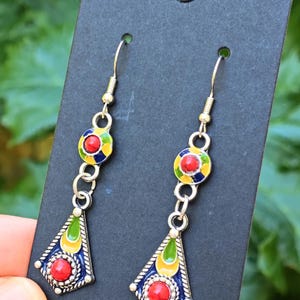 Puede incluir: Un par de pendientes colgantes con un diseño geométrico colorido. Cada pendiente presenta una piedra roja redonda engastada en un marco de metal plateado, con detalles de esmalte azul, amarillo y verde. Los pendientes están suspendidos de un gancho simple.
