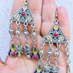 Puede incluir: Pendientes colgantes plateados con una parte superior triangular y abalorios colgantes. Los pendientes presentan detalles de esmalte rojo, azul, amarillo y verde. Los abalorios incluyen pequeñas cuentas redondas y colgantes decorativos.